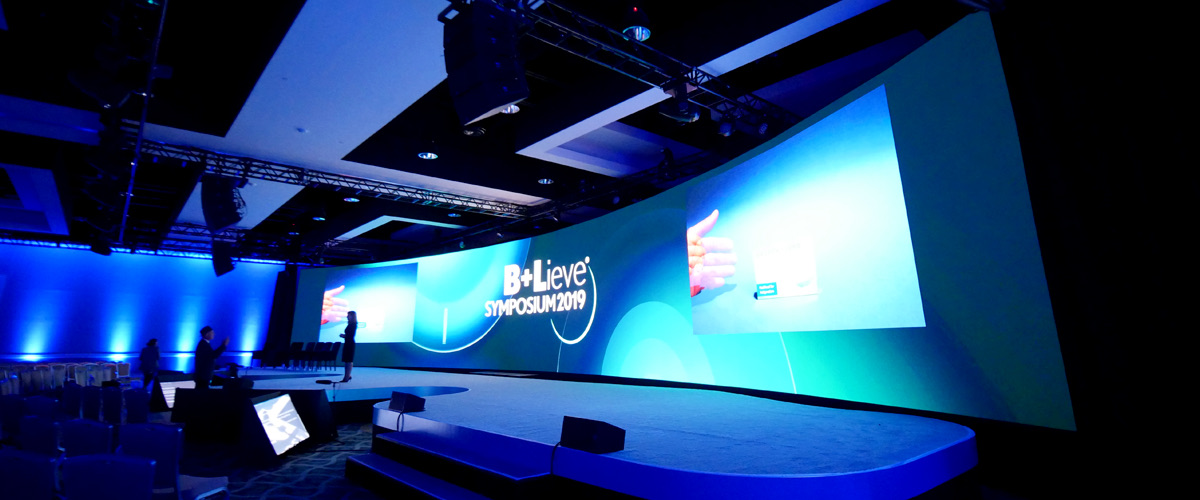 Corporate keynote stage with a wide blended projection screen and blue lighting