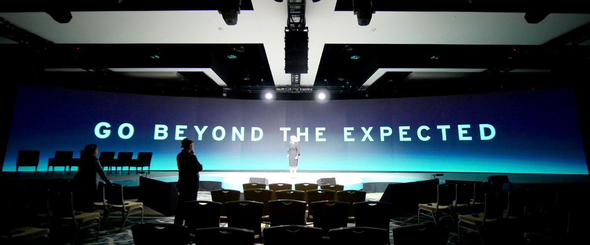 A presenter on stage in front of a wide curved screen reading Go Beyond the Expected, two people watching from rows of empty chairs